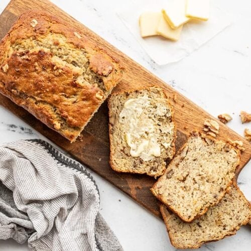 A loaf of yogurt banana bread on a wooden cutting board, a few slices scattered, one smeared with butter