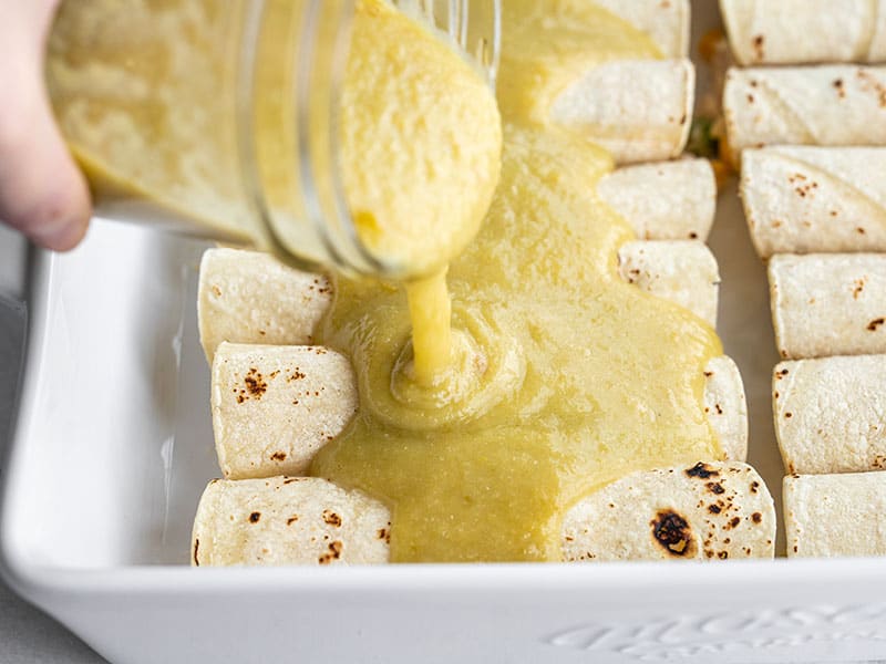 Green Chile Enchilada Sauce being poured from a jar onto a pan of rolled enchiladas