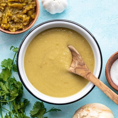 A wooden spoon in a bowl of green chile enchilada sauce with cilantro and diced green chiles on the side