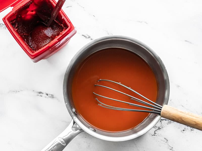 Gochujang broth in a small sauce pot with a whisk, tub of gochujang on the side