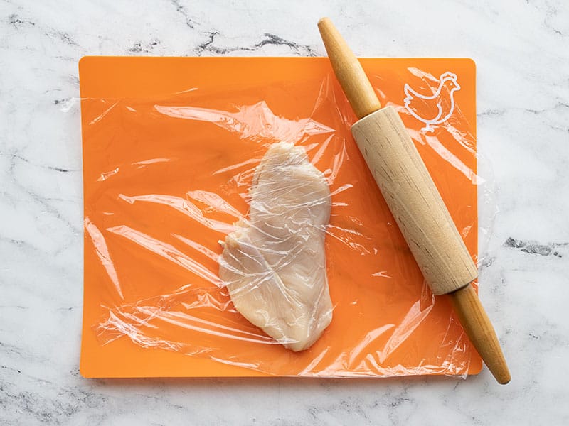 Chicken breast on a cutting board covered with plastic, pounded with a rolling pin