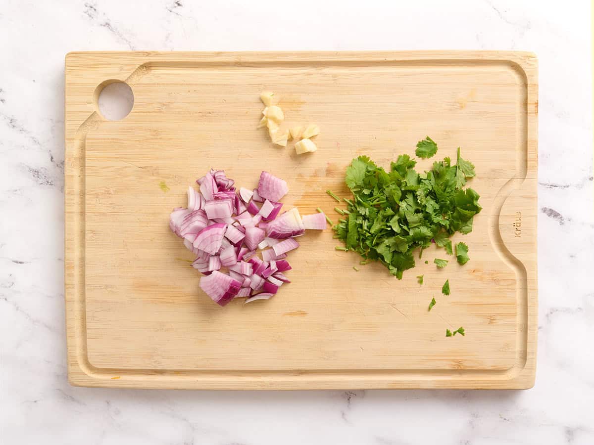 Roughly chopped red onion, garlic and cilantro on a wooden cutting board.