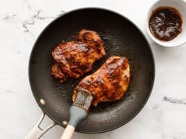 BBQ sauce being brushed onto chicken in a skillet