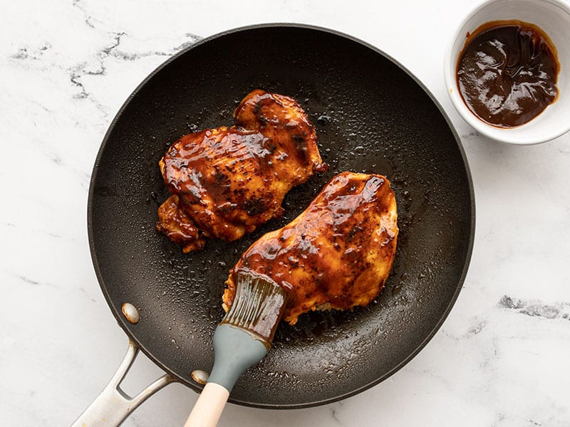 BBQ sauce being brushed onto chicken in a skillet