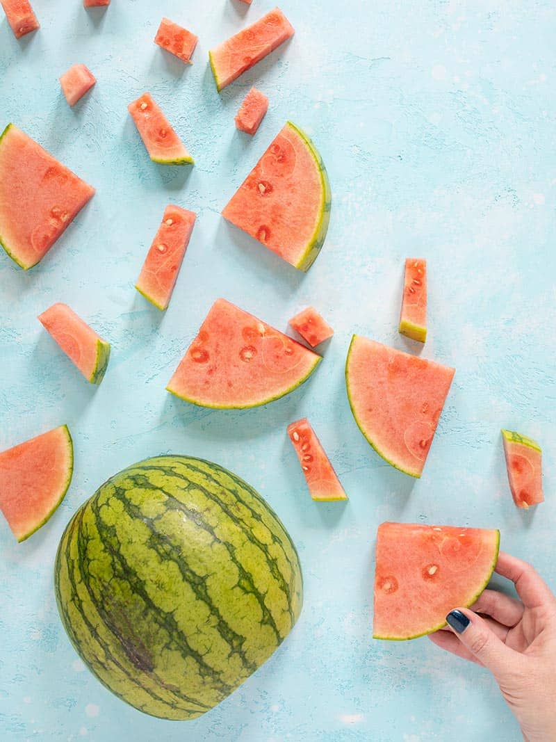 A watermelon cut into several different shape and sized pieces, a hand grabbing a wedge