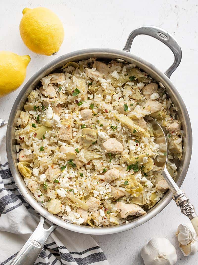 One Pot Lemon Artichoke Chicken and Rice in the skillet with lemon and garlic on the side
