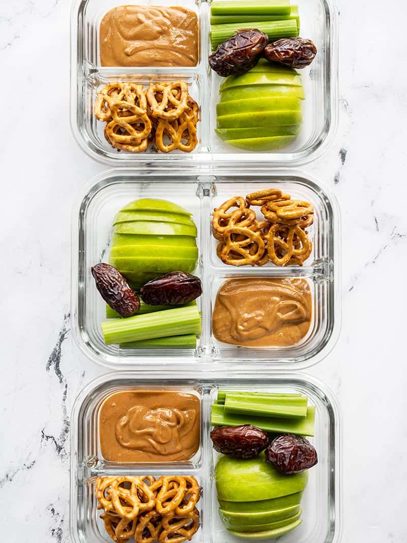 Three glass containers lined up with peanut butter apples, celery, pretzels and dates.