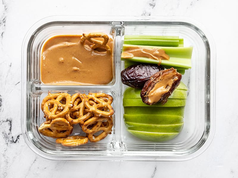 A single peanut butter lunch box with peanut butter on a Medjool date and a pretzel dipping into the peanut butter