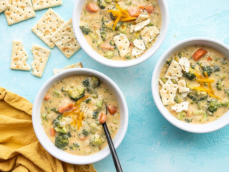 Three bowls of broccoli cheddar soup with saltines on the side