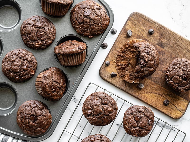 Chocolate banana muffins in the muffin tin, on a cooling rack, and on a cutting board