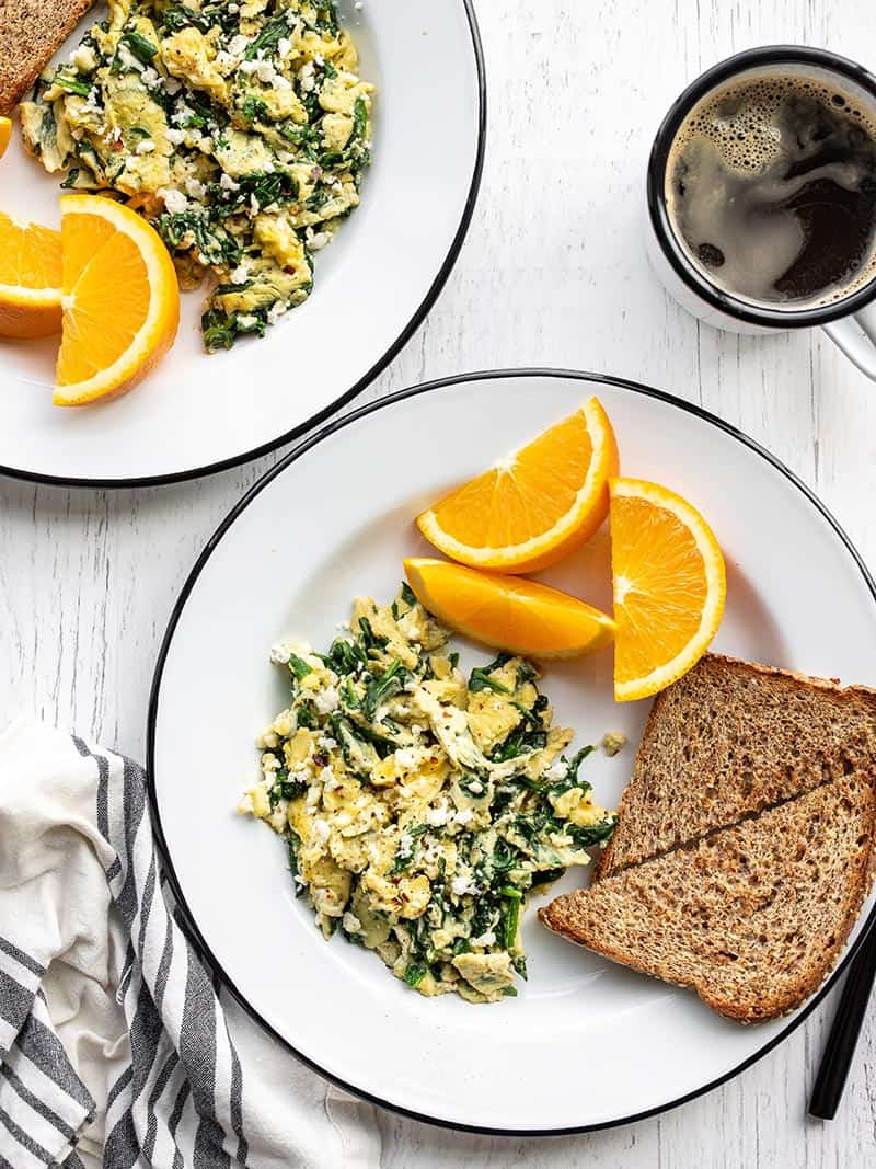 Two plates with scrambled eggs with spinach and feta, toast, and orange slices