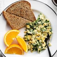 A plate full of scrambled eggs with spinach and feta, toast, and orange slices