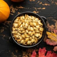 Roasted pumpkin seeds in a small black ceramic bowl with pumpkins and leaves on the sides
