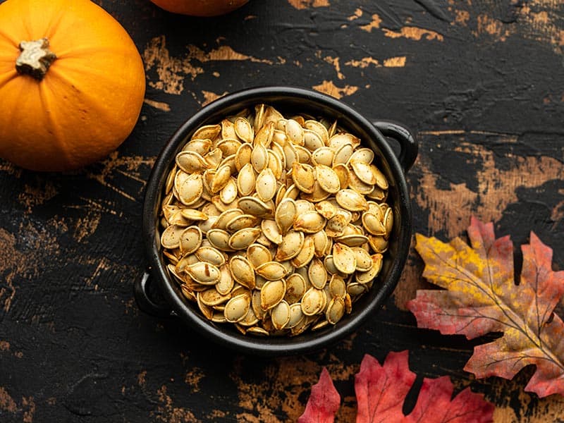 Roasted pumpkin seeds in a small black ceramic bowl with pumpkins and leaves on the sides