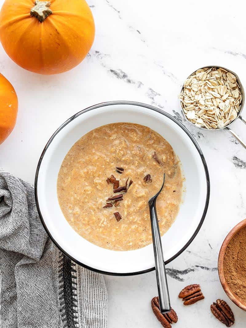 Overhead view of a bowl of overnight pumpkin pie oats with pumpkins, oats, and pecans on the side