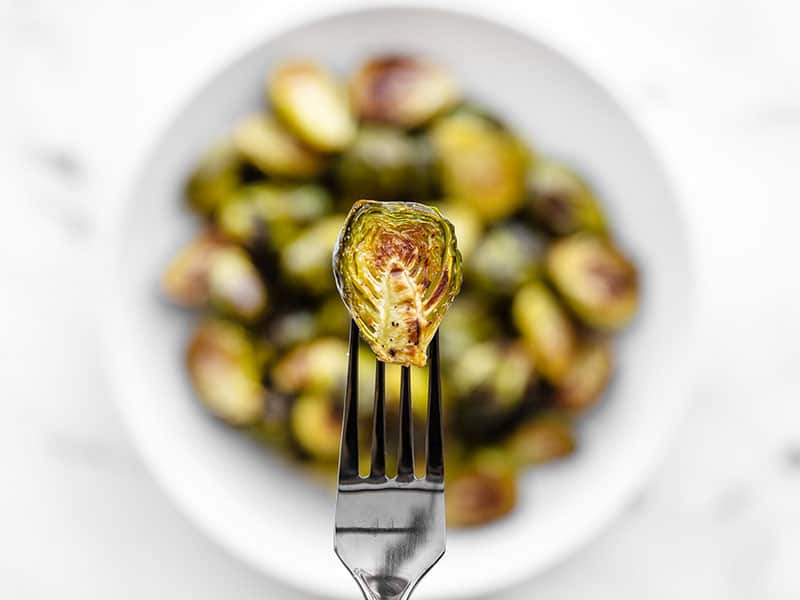 Close up of a roasted Brussels sprout on a fork with the bowl in the background