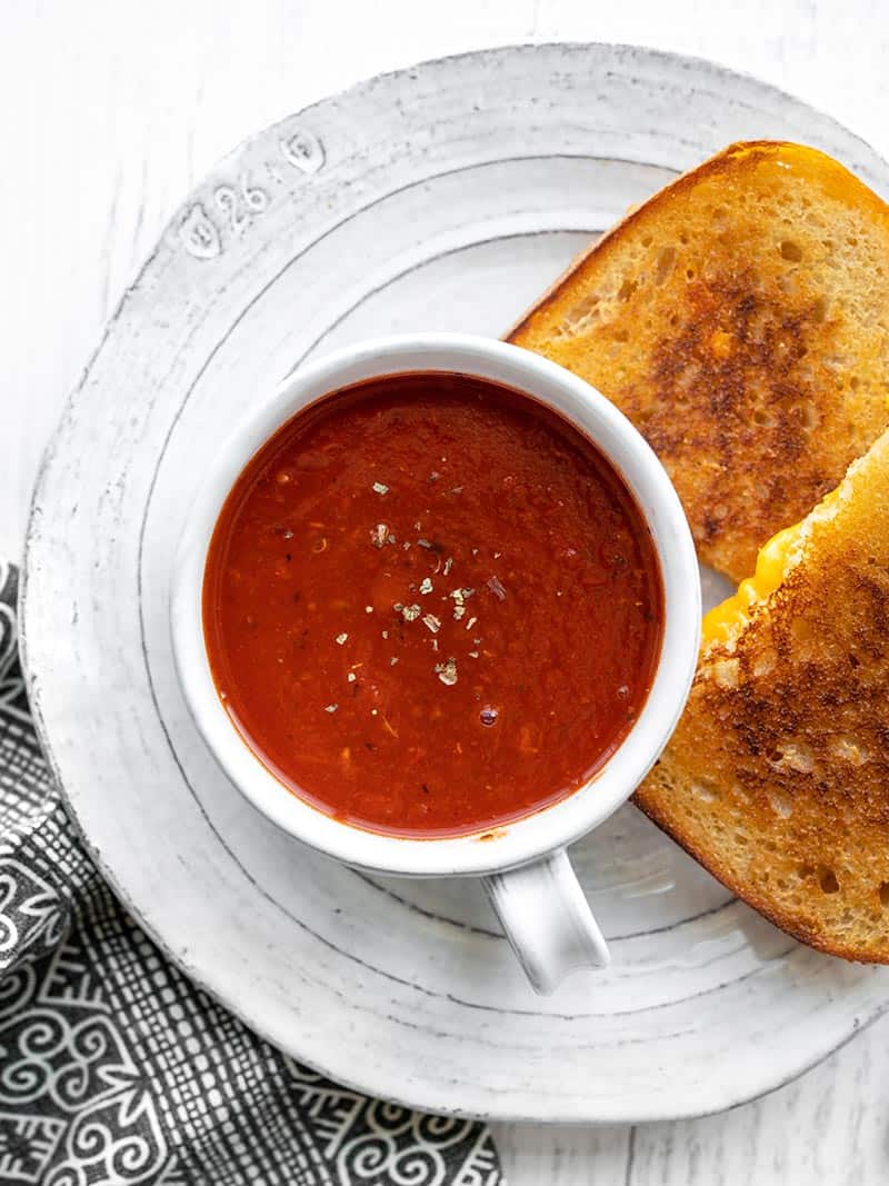 A mug of tomato herb soup on a plate with a grilled cheese