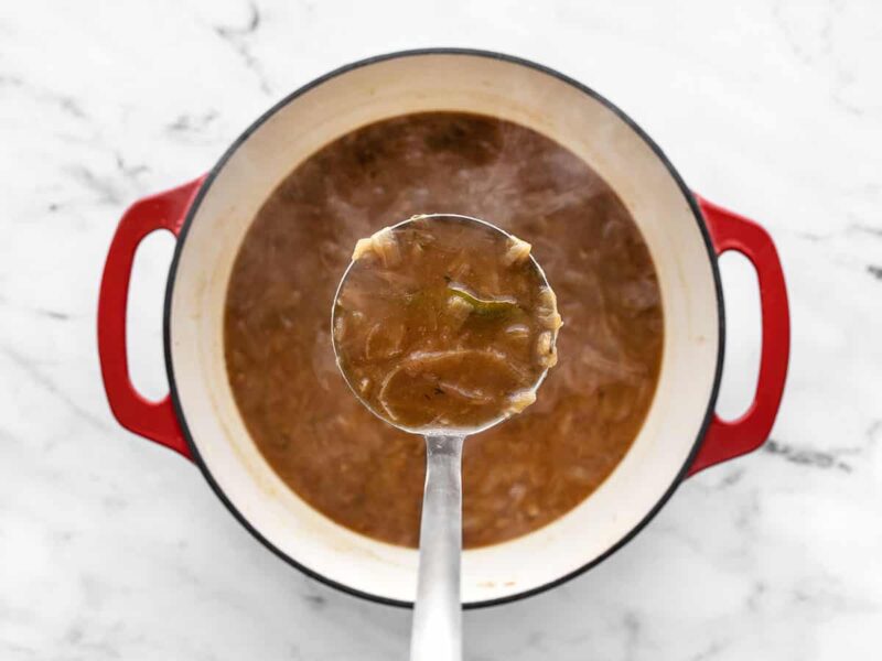 A pot of French Onion Soup with a ladle full held close to the camera