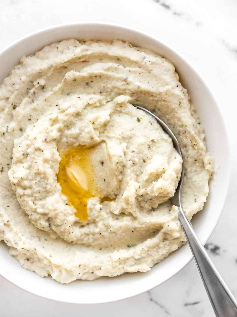 Mashed cauliflower in a bowl with a spoon and a pat of melted butter in the middle