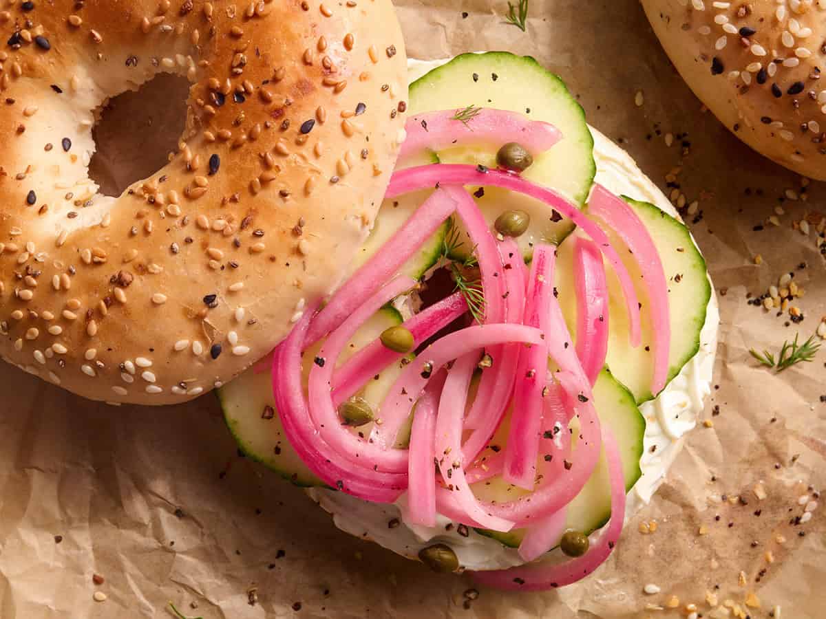Overhead view of a cream cheese bagel with sliced cucumber and pickled red onions.