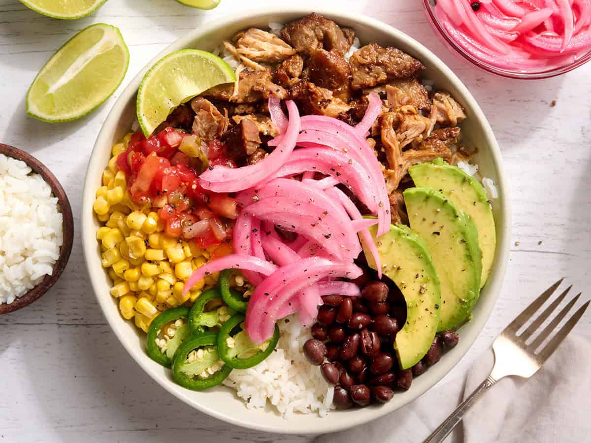 A taco bowl with pulled pork, sliced avocado, jalapeno, corn, rice, salsa, and pickled red onions.