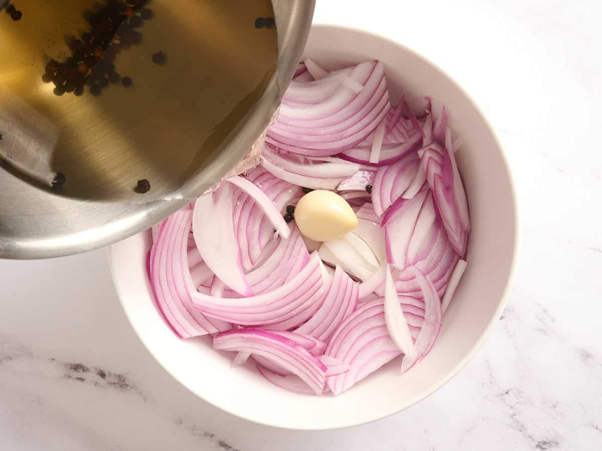 A hot vinegar pickle mixture being poured over sliced red onions and garlic in a bowl.