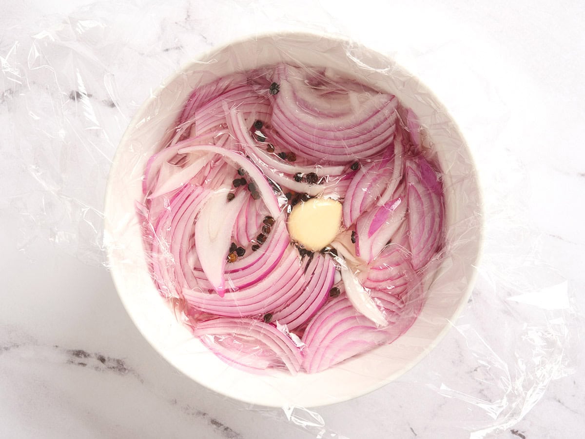 Plastic wrap placed over a bowl of sliced red onions, pepper corns and garlic.