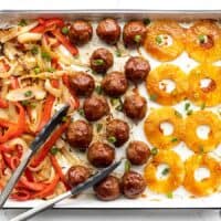 Overhead view of sheet pan with bbq meatballs, pineapple, peppers, and onions, tongs on the side