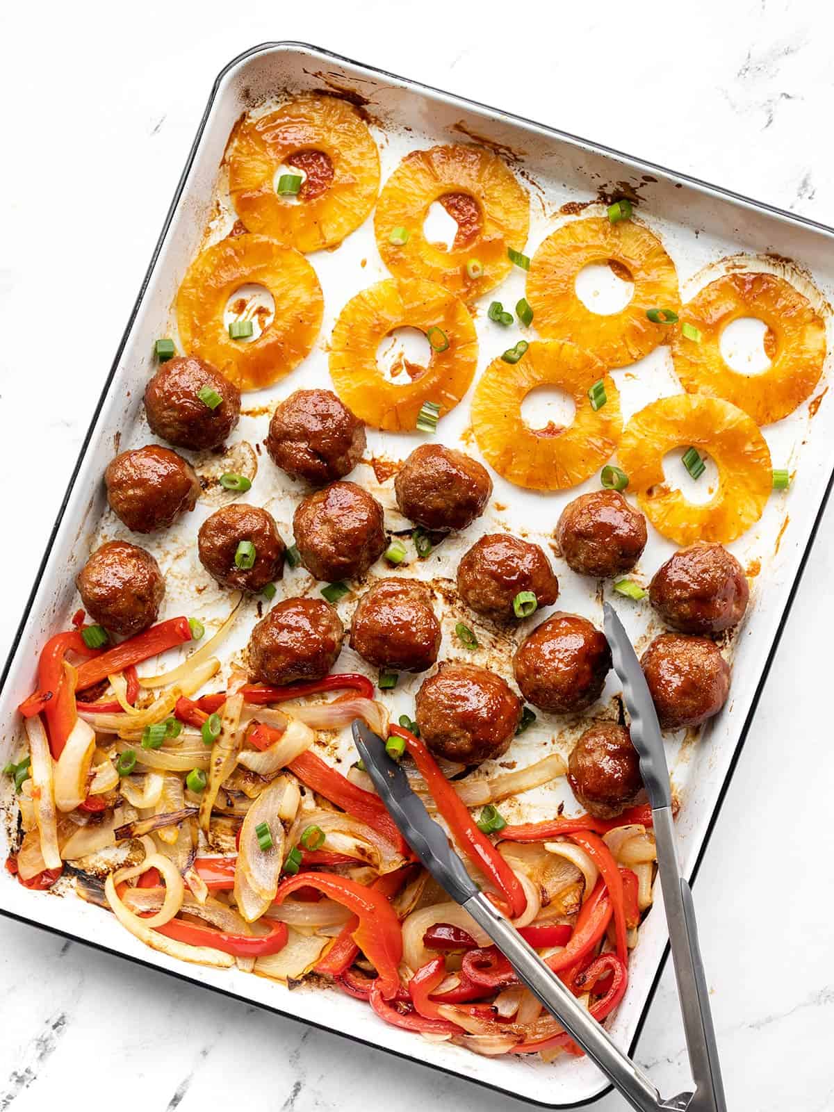 Sheet pan bbq meatballs on the pan with pineapple, peppers, and onions. Tongs resting on the side.