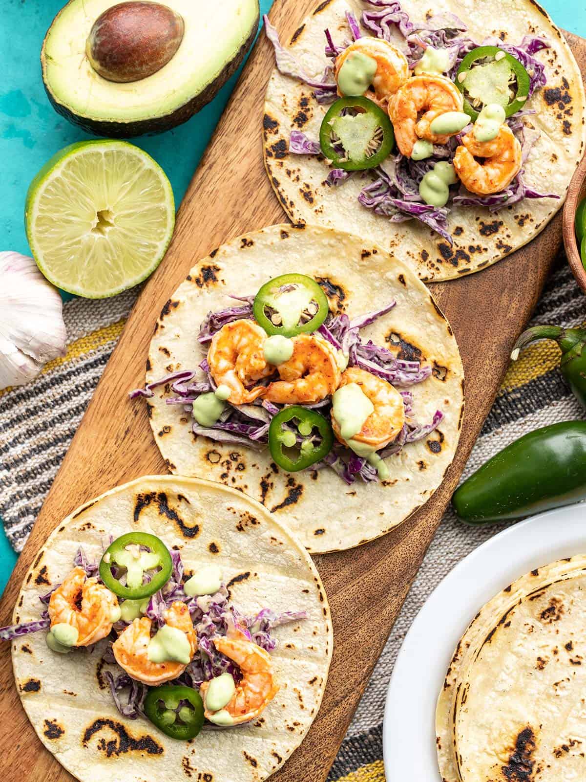 Three Tajín Shrimp Tacos open faced on a wooden board, ingredients on the sides
