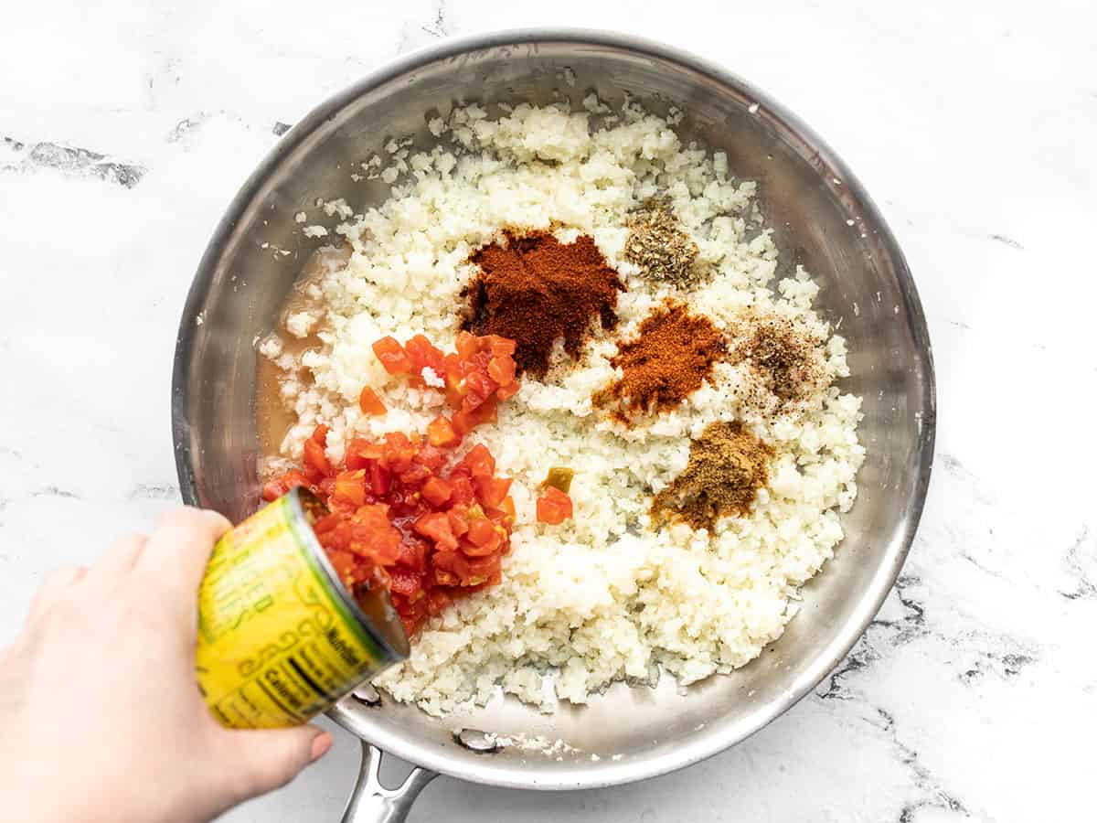 Tomatoes being poured into the skillet with cauliflower rice and spices