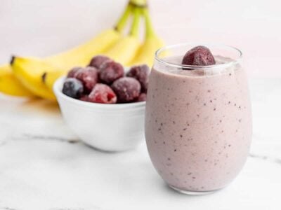 A glass filled with a cherry almond smoothie, with bananas and a bowl of frozen cherries in the back