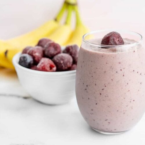 A glass filled with a cherry almond smoothie, with bananas and a bowl of frozen cherries in the back