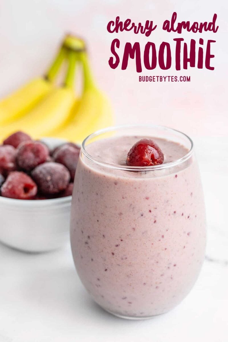 A glass filled with Cherry Almond Smoothie, banana and a bowl of frozen cherries in the back