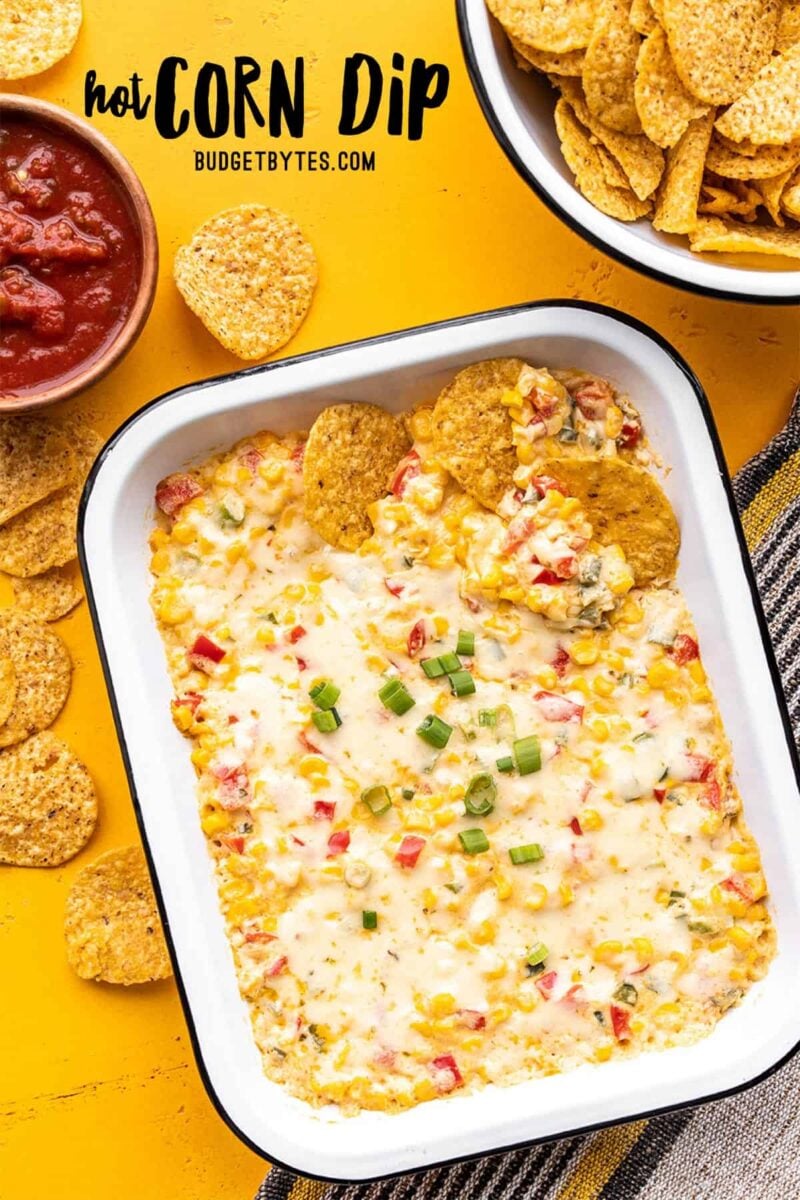 A rectangular casserole dish of hot corn dip with chips and salsa on the side