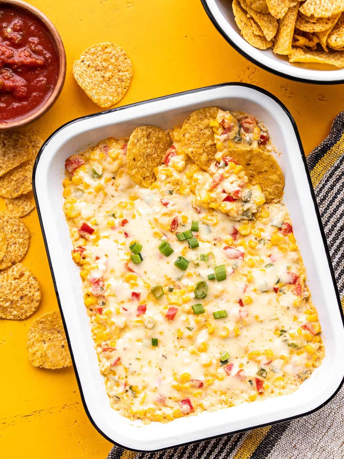 A rectangular baking dish full of hot corn dip with chips and salsa on the sides