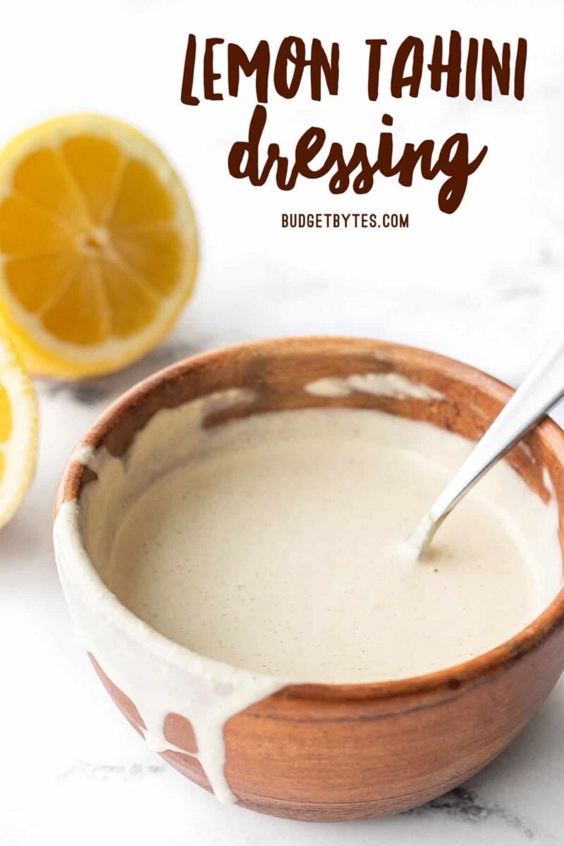 Lemon tahini dressing in a wooden bowl, some dripping off the side. Lemons in the back.