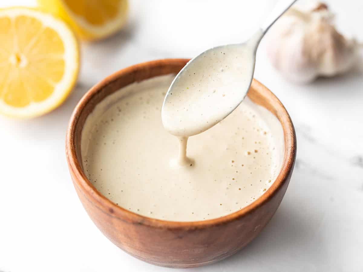 Lemon tahini dressing dripping off a spoon into a wooden bowl. Lemons in the background