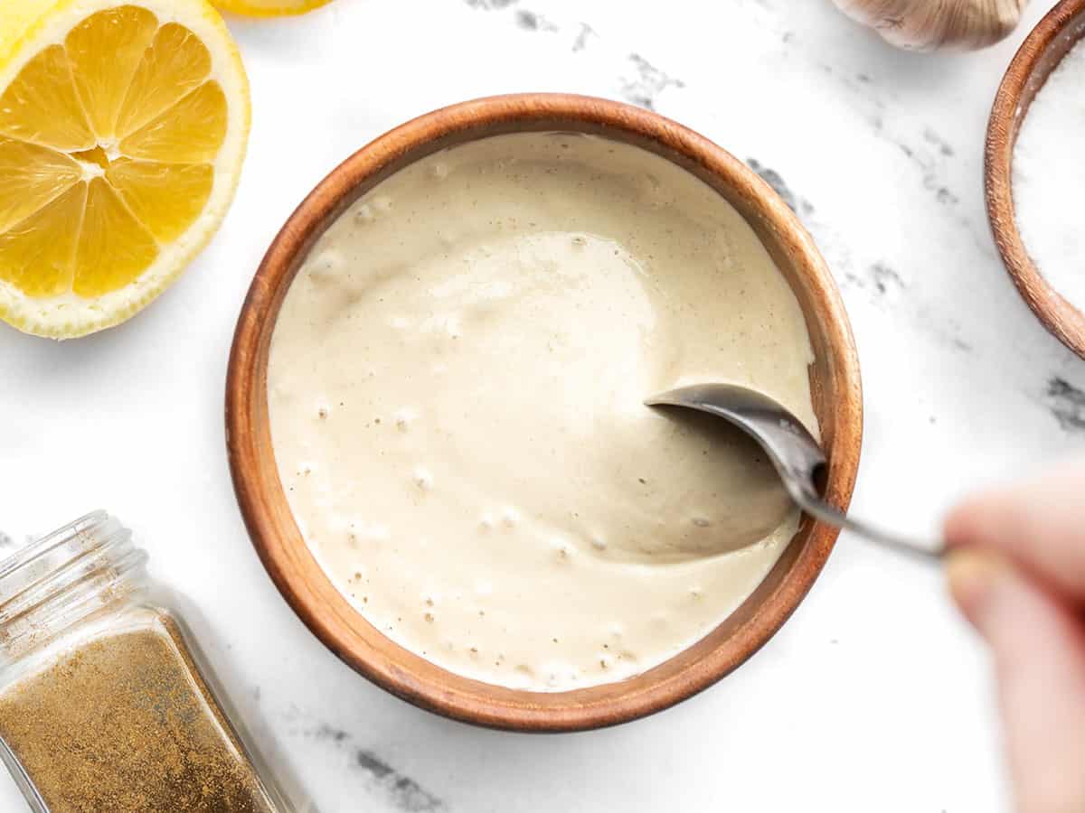 A spoon stirring the thick lemon tahini dressing in a wooden bowl, ingredients on the sides
