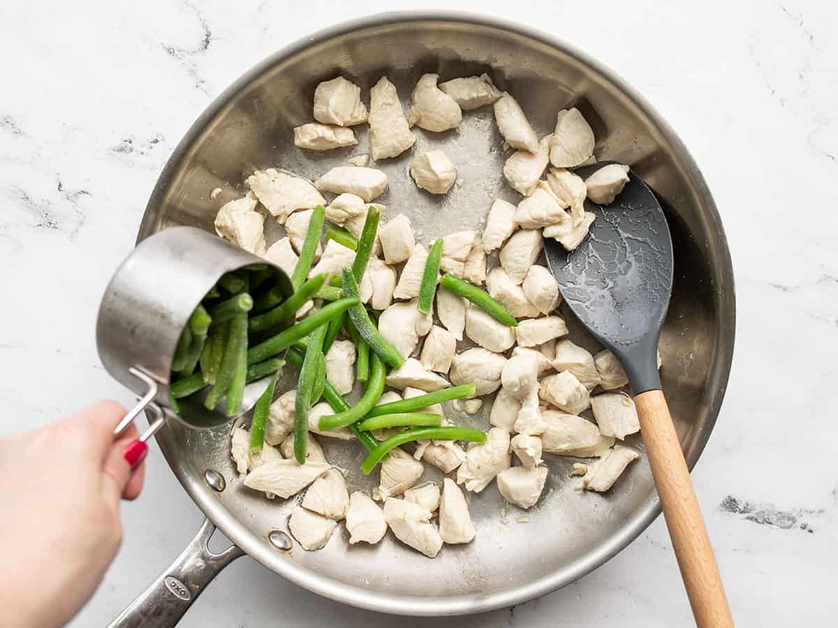 Cooked chicken in the skillet, frozen green beans being poured in