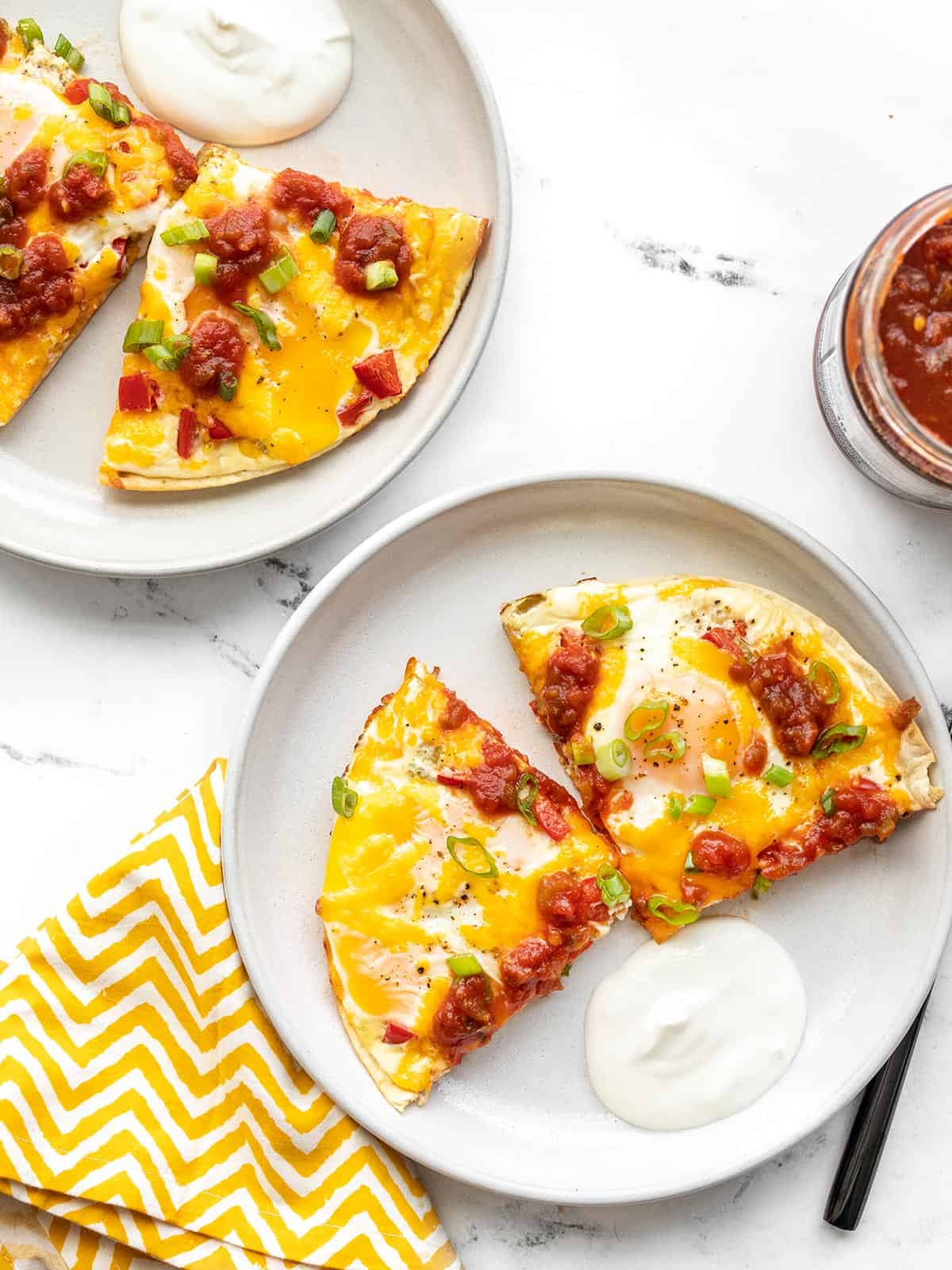 Two plates with southwest tortilla baked eggs and sour cream, a jar of salsa on the side