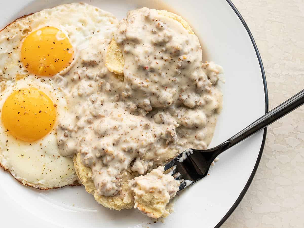 A fork taking a bite of biscuits and gravy off a plate with eggs