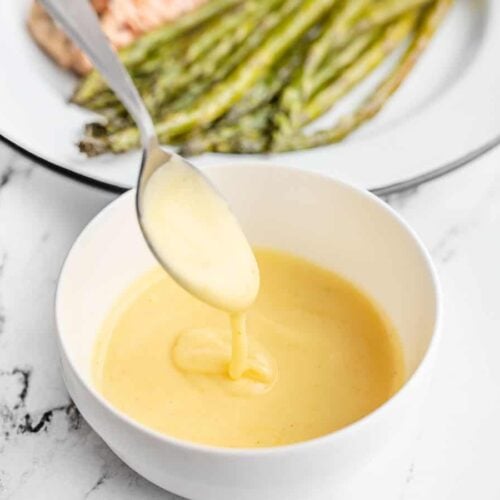 hollandaise sauce dripping off a spoon into a bowl in front of a plate of asparagus