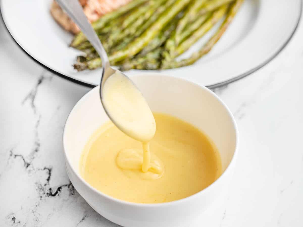 hollandaise sauce dripping off a spoon into a bowl in front of a plate of asparagus