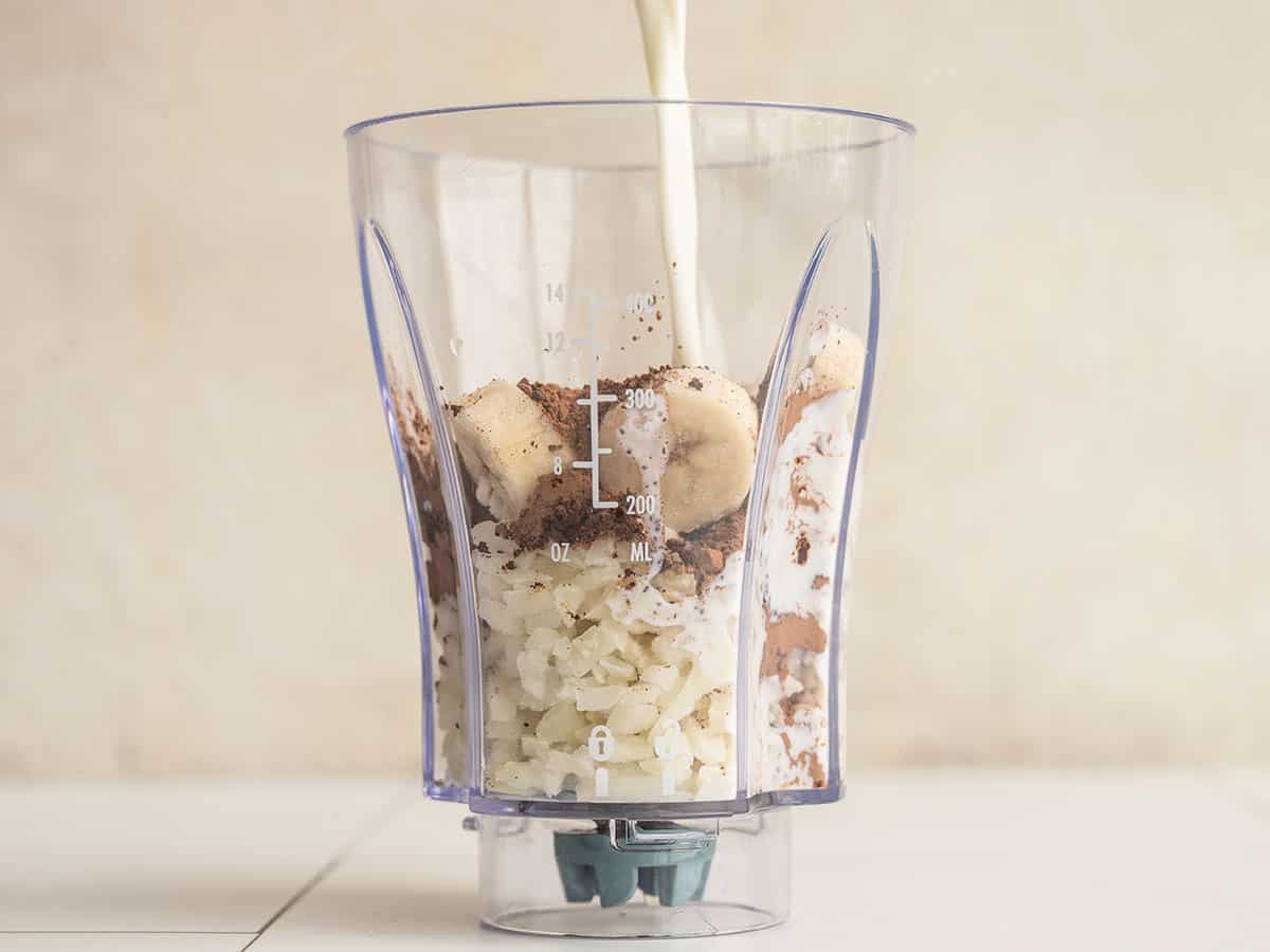 Milk being poured into a blender with cauliflower, bananas, cocoa, and instant coffee