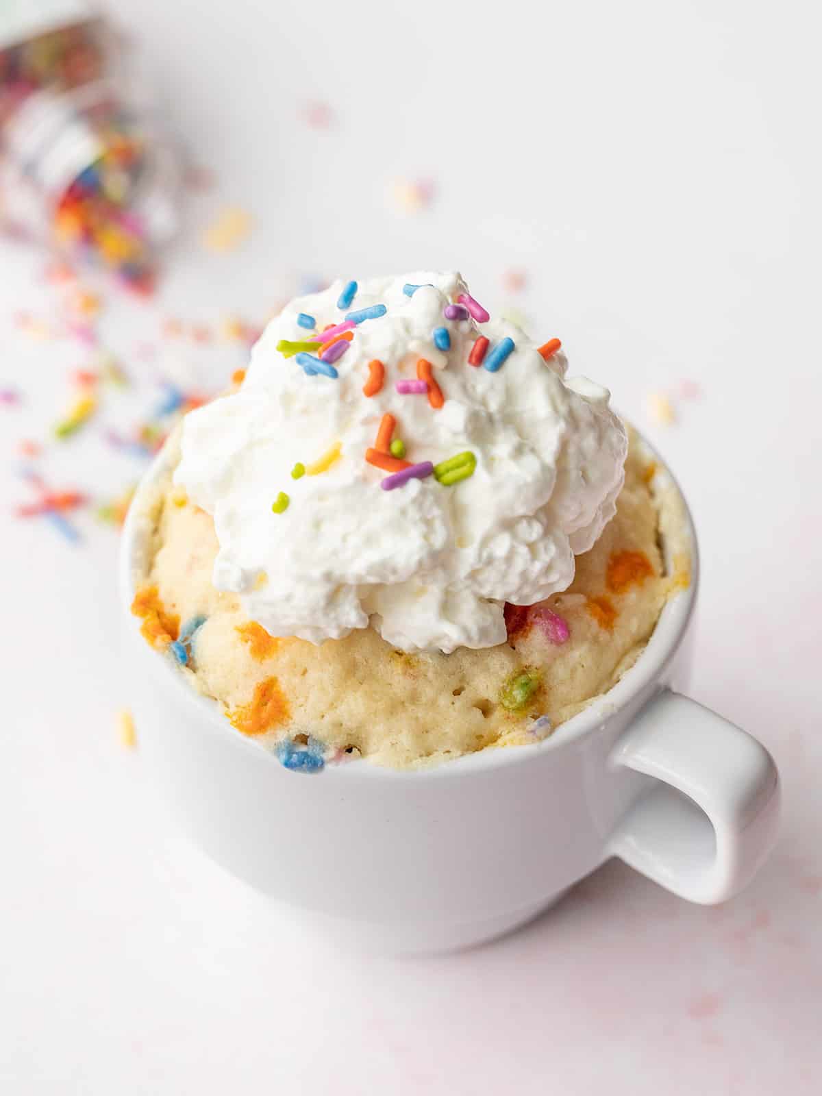 A funfetti mug cake topped with whipped cream, sprinkles spilled in the back