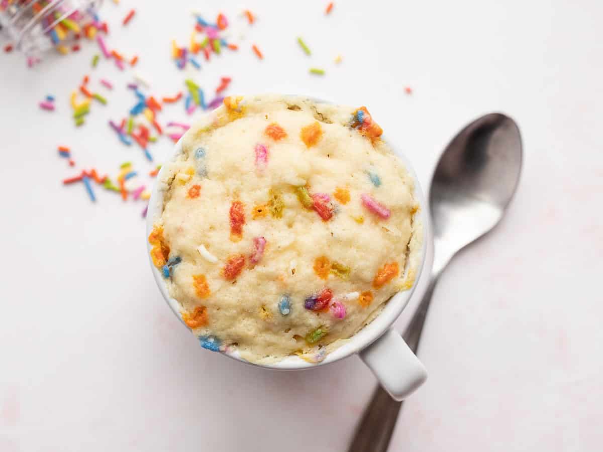 Overhead view of an unfrosted funfetti mug cake with a spoon on the side and sprinkles in the background