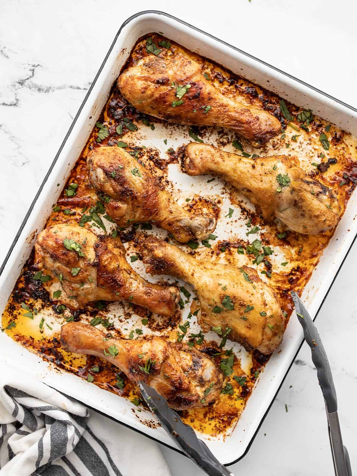 baked chicken drumsticks on a baking sheet with tongs, garnished with parsley