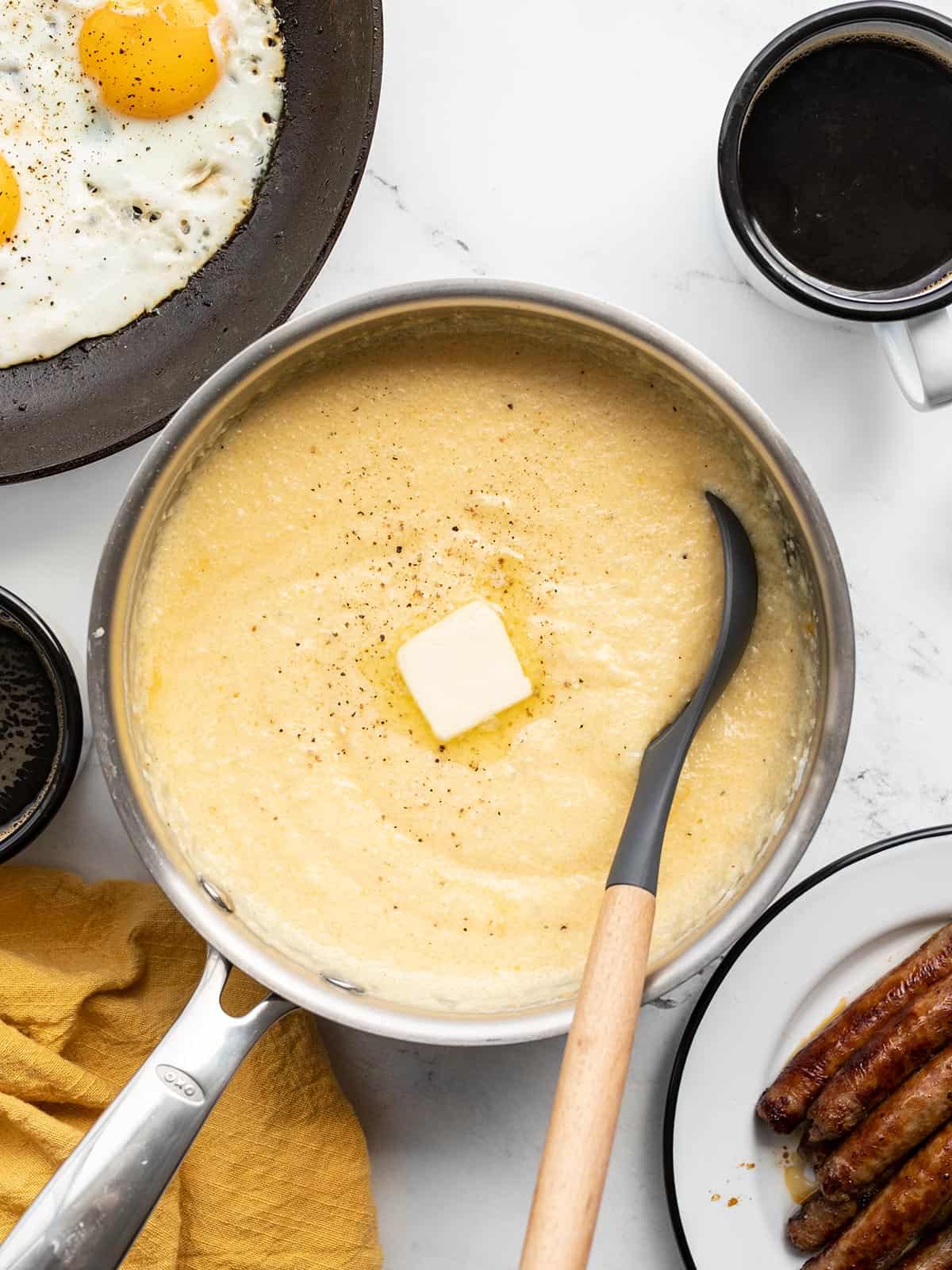 A pot full of cheese grits with a pat of butter, other breakfast items on the sides
