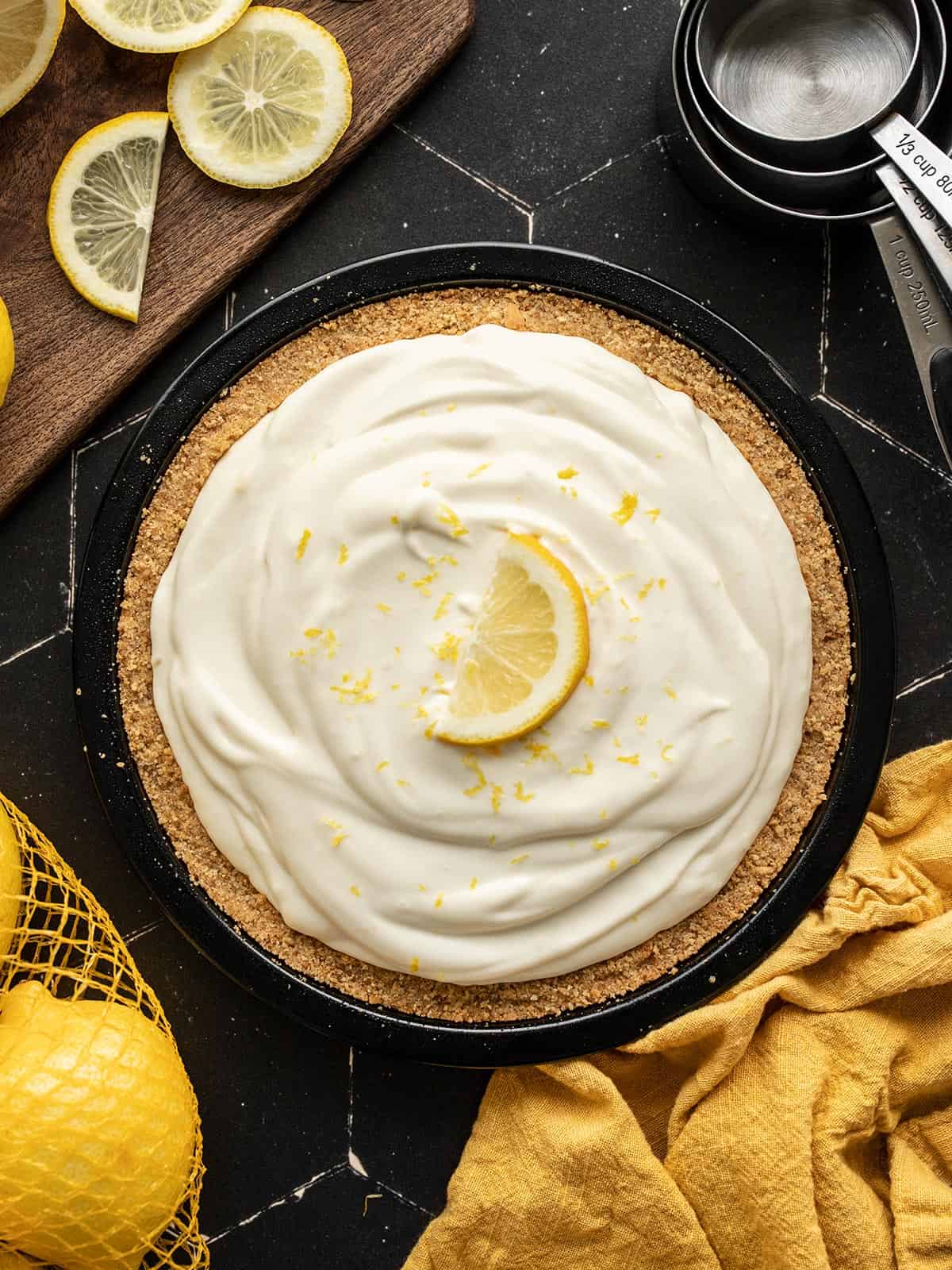 Overhead view of a lemon cream pie on black tile with lemons on the side